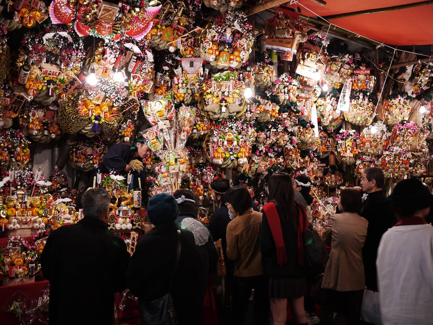 Walls of decorated kumade rakes lit up at a Tori-no-ichi stall after dark