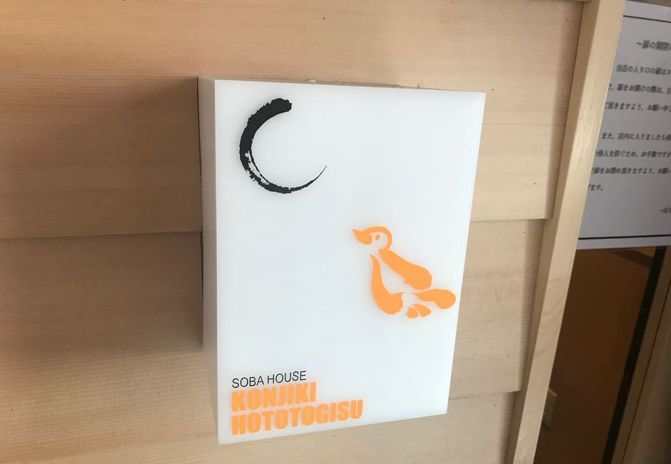 The Soba House Konjiki Hototogisu sign with its orange bird logo on a pale wooden wall in Shinjuku